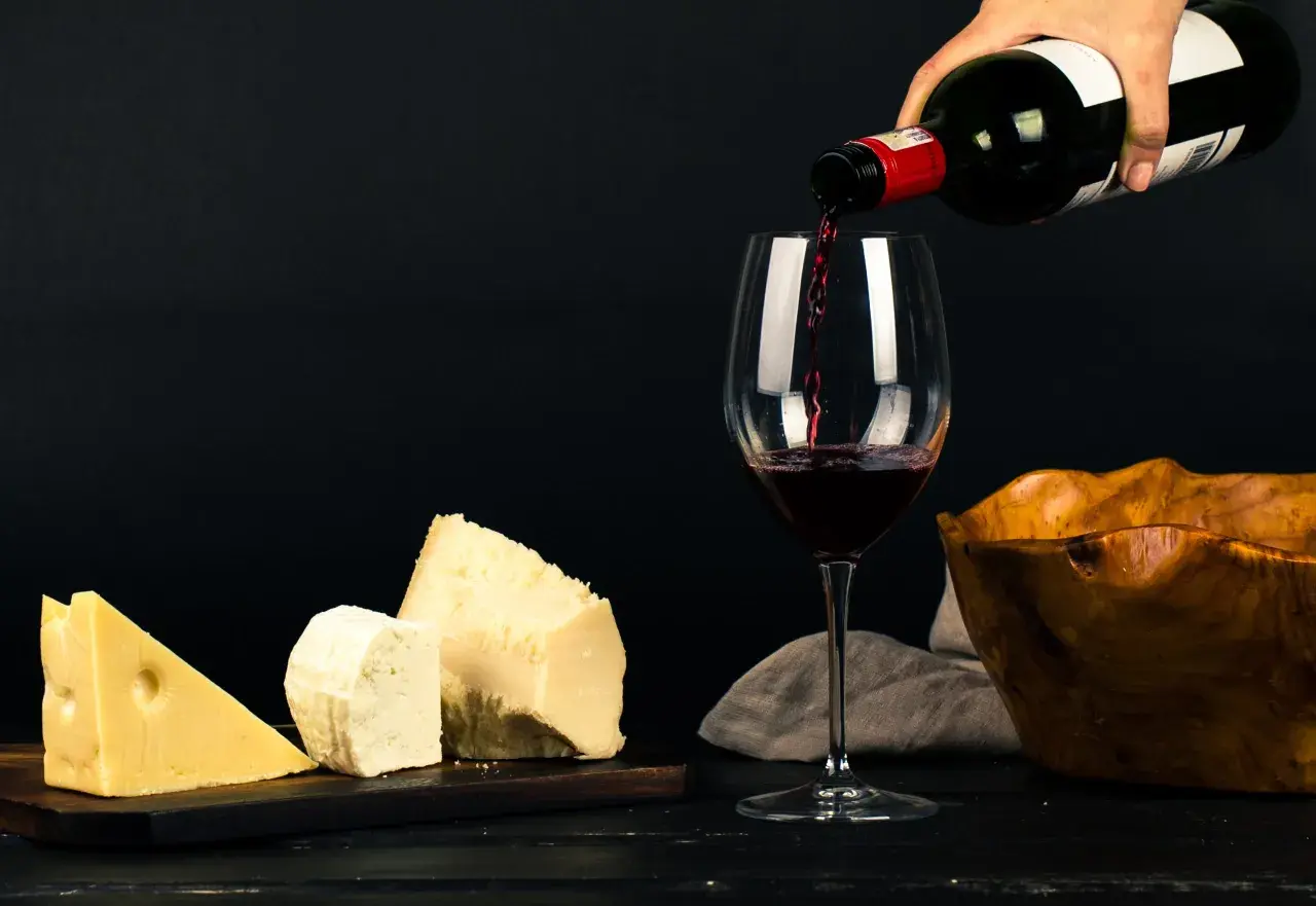 Un verre se remplit de vin rouge, à côté de morceaux de fromage et d'un bol en bois.
