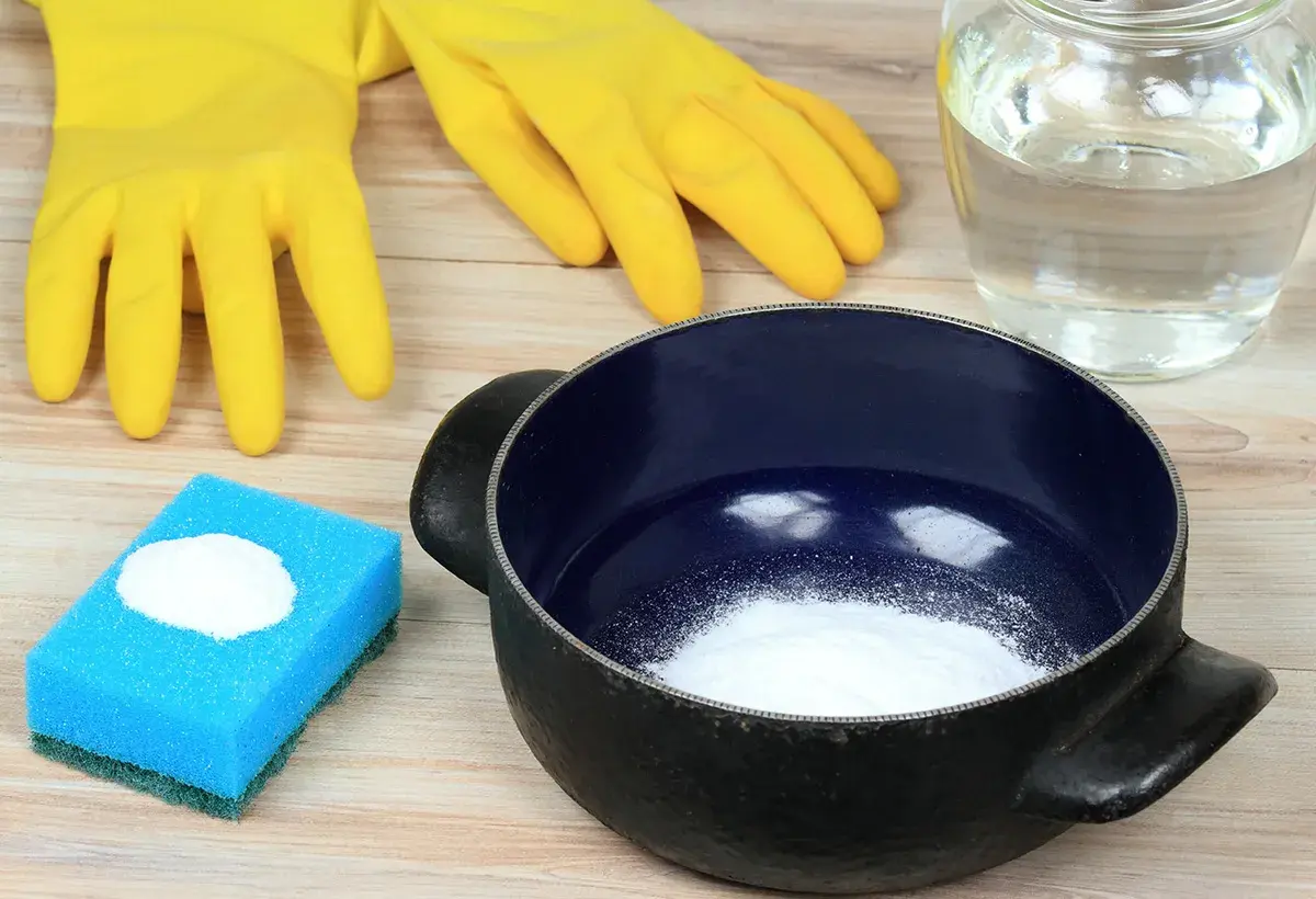 Gants jaunes, éponge bleue avec poudre blanche, casserole noire et un bocal rempli de liquide. Idéal pour savoir comment enlever le cramer dans une casserole sans bicarbonate.