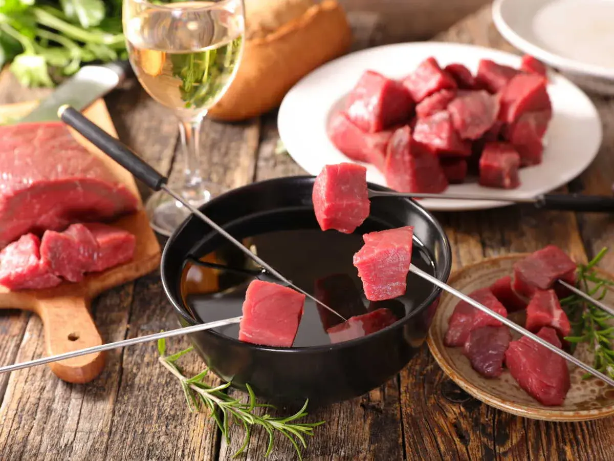 Préparer une fondue bourguignonne : des dés de bœuf crus sur des brochettes, prêts à être plongés dans l'huile chaude. Un verre de vin blanc à côté.
