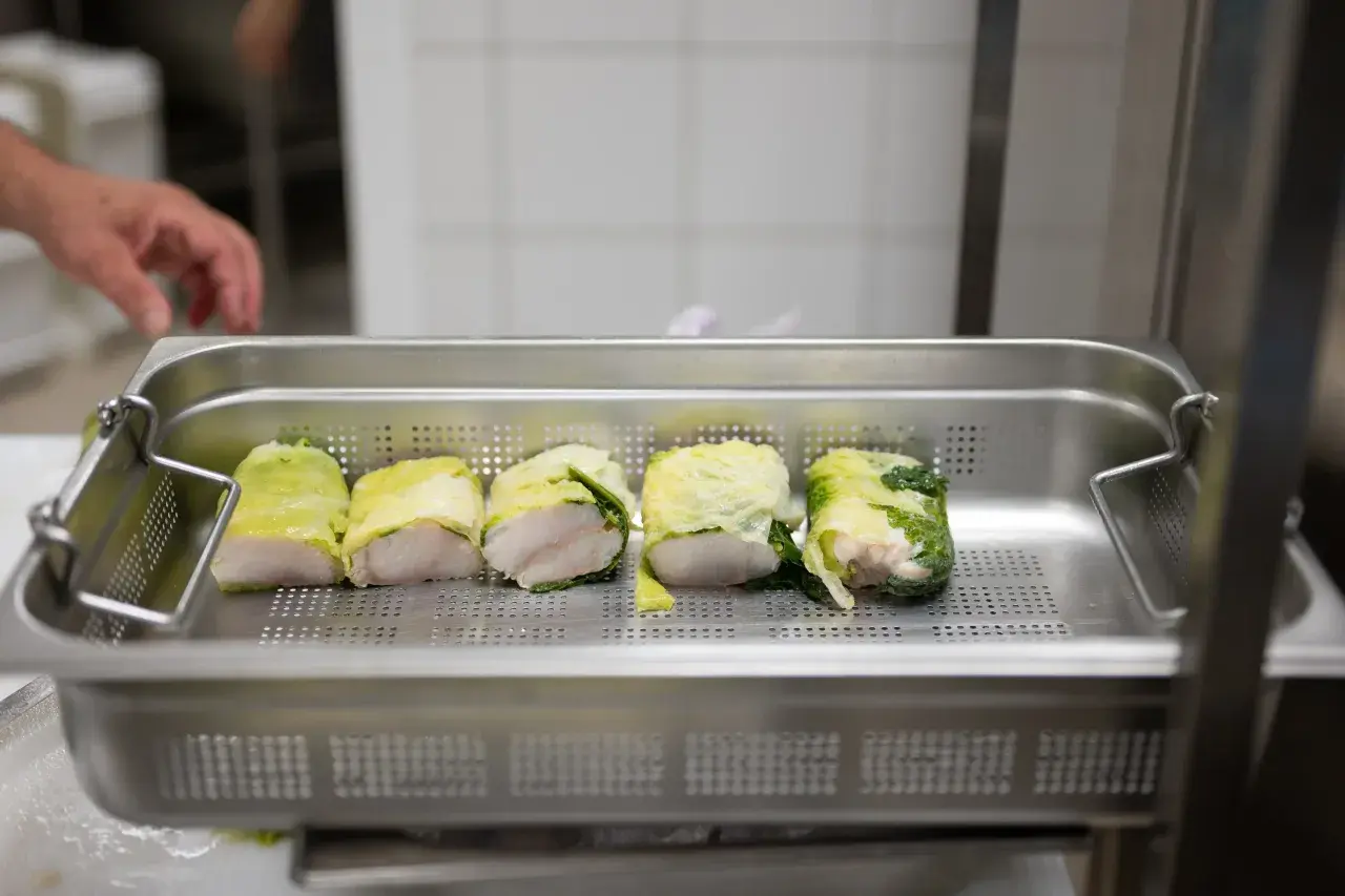 Cinq rouleaux de poisson enroulés dans des feuilles de chou, prêts pour la cuisson. Une main est visible en arrière-plan, préparant ce plat pour la journée internationale des cuisiniers.