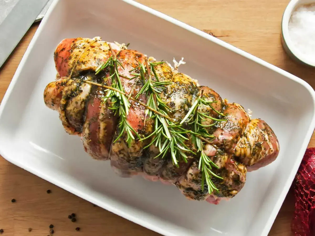 Un rôti de porc rosé, assaisonné et garni de romarin frais, prêt à être cuit dans un plat blanc.