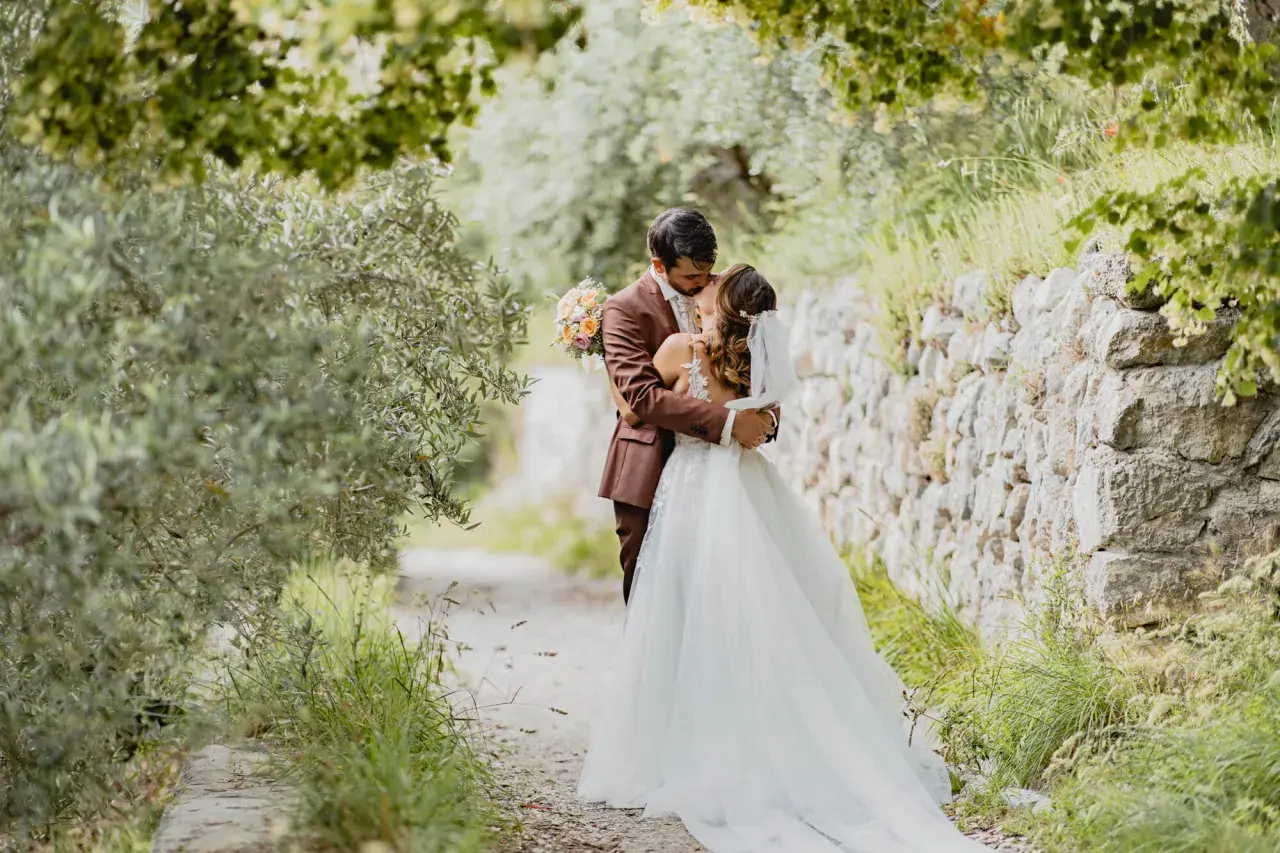 Un couple s'embrasse tendrement lors de leur mariage de 50 personnes, entouré par la nature verdoyante et un mur de pierres anciennes.