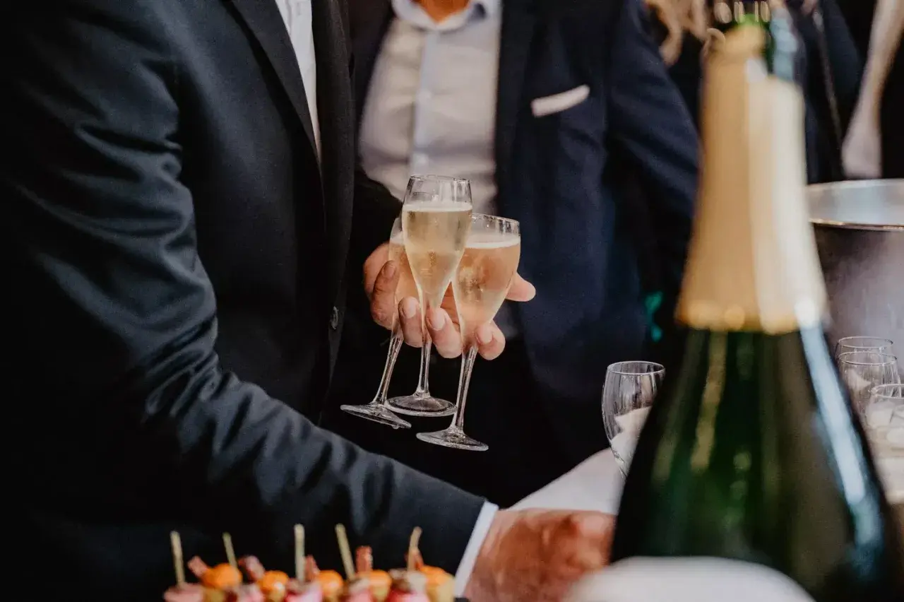 Des hommes en costume portent des flûtes de champagne. Une bouteille de champagne est au premier plan, suggérant un vin d'honneur original.
