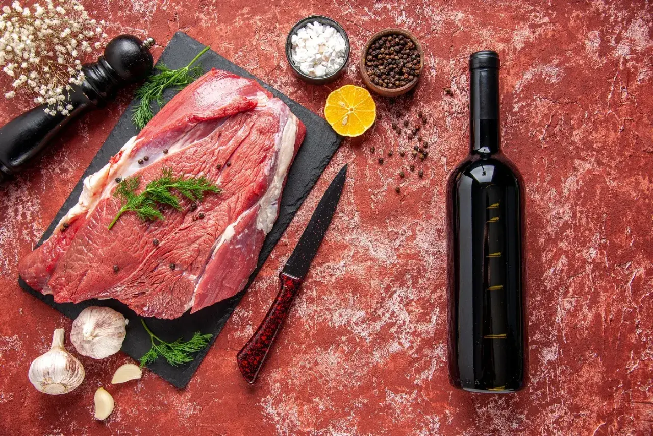 Un morceau de viande crue, prêt pour un repas savoureux avec un vin rouge. Des épices, de l'ail et une tranche de citron complètent la scène.