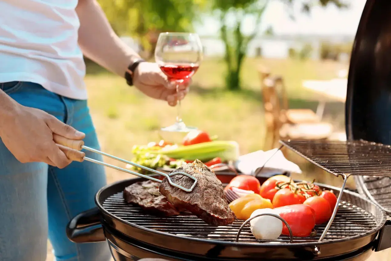 Un homme savoure un verre de vin rouge tout en préparant un barbecue avec des steaks et des légumes.