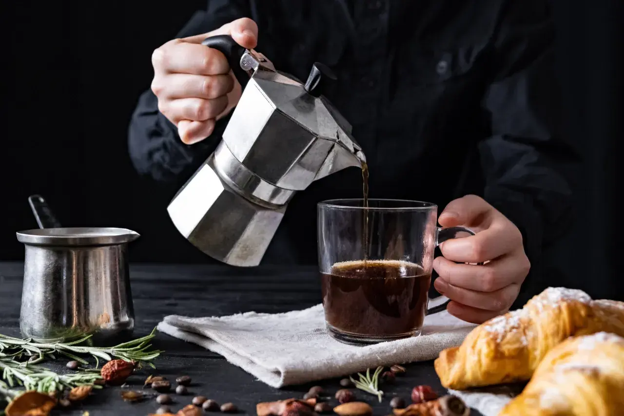 Cafetière italienne inox versant du café dans une tasse. Un petit pot en métal et des croissants sont sur la table.
