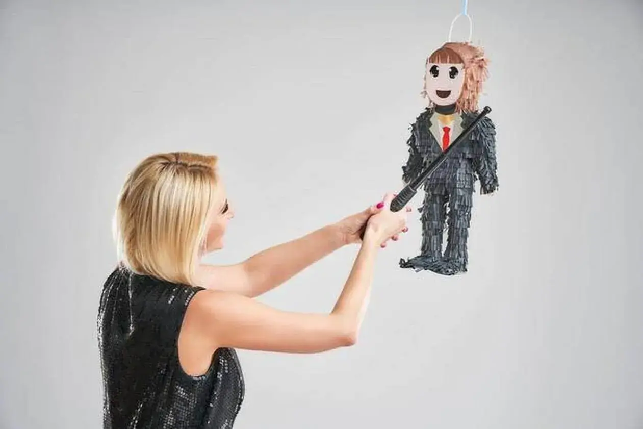 Une femme en robe à paillettes s'apprête à frapper une piñata en forme d'homme en costume, symbole d'une fête de divorce réussie.