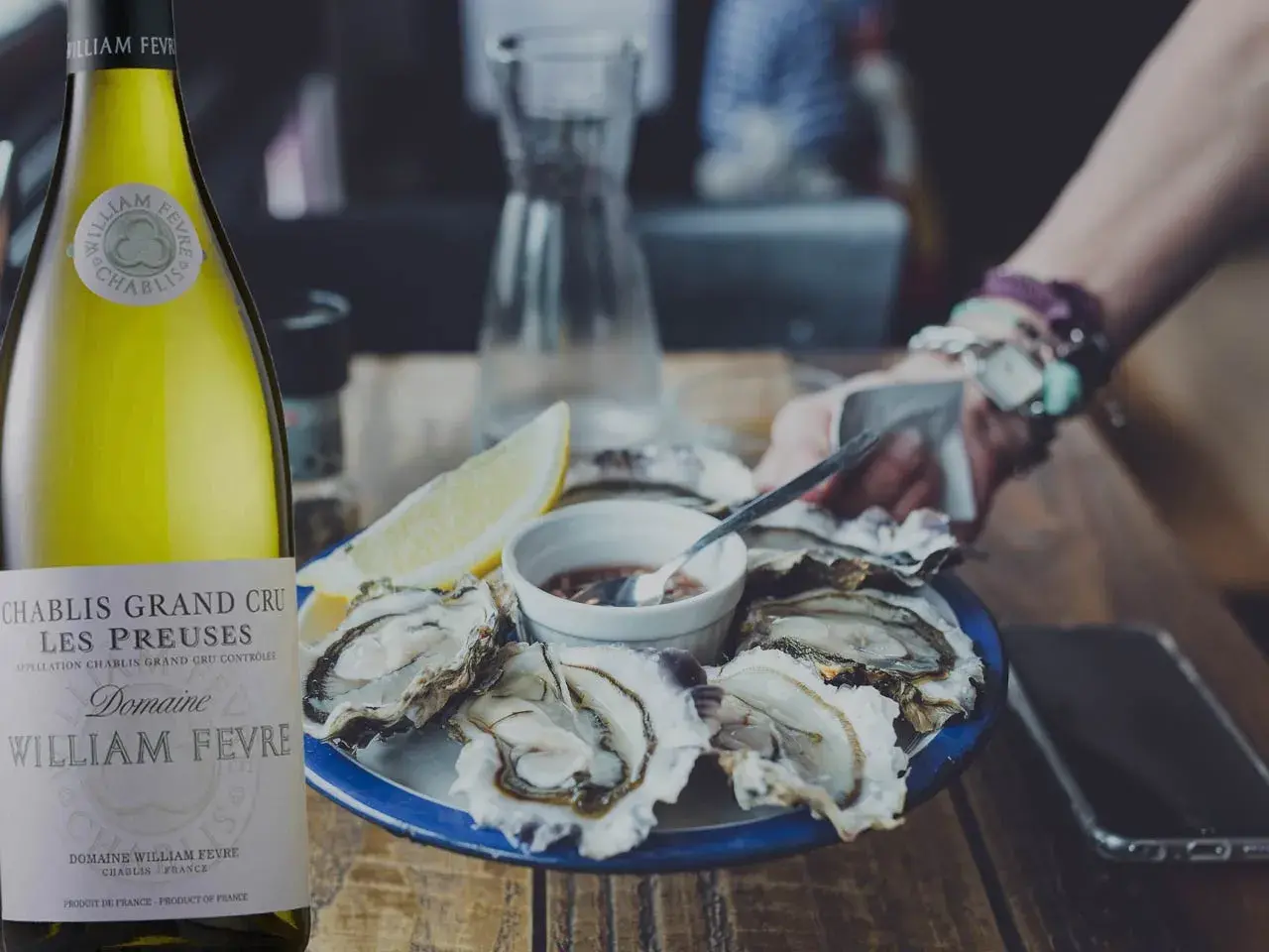 Une bouteille de Chablis Grand Cru William Fèvre, parfaite pour savoir quoi boire avec des huîtres. Une assiette d'huîtres fraîches et un quartier de citron complètent la scène.
