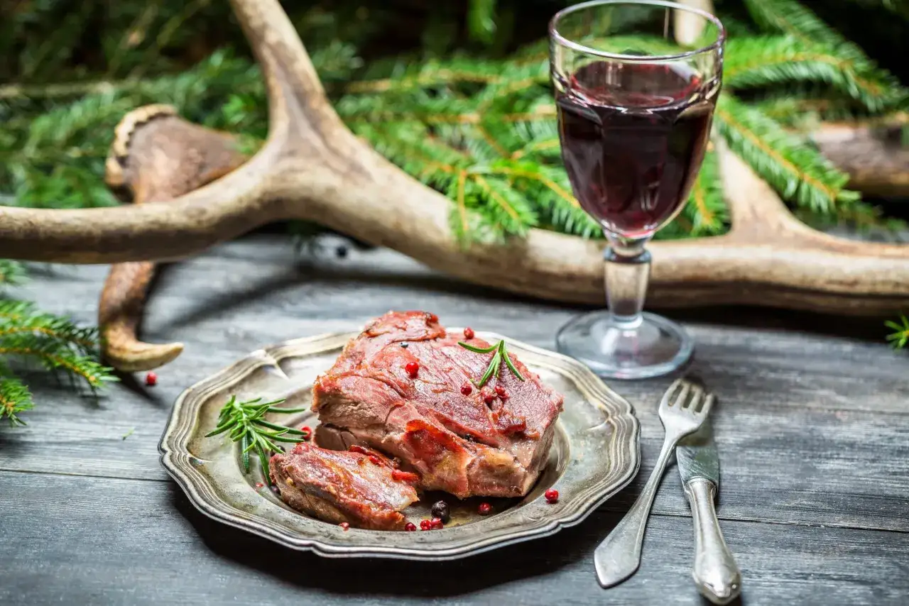 Un plat de viande rouge, garni de romarin et de baies roses, prêt à être dégusté avec un verre de vin pour civet. Des bois de cerf et des branches de sapin en arrière-plan.