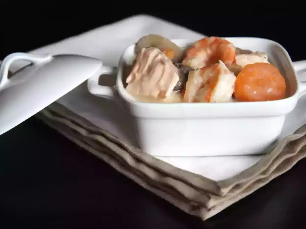 Un plat de blanquette de saumon et crevettes, servi dans une cocotte blanche avec une sauce onctueuse au vin blanc.