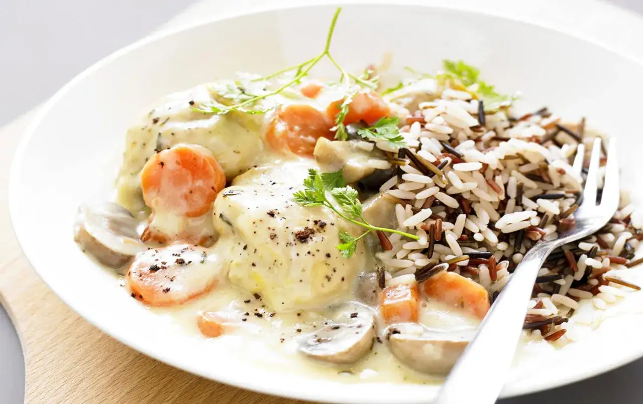 Un plat de blanquette de poulet crémeuse avec des carottes, des champignons et un accompagnement de riz sauvage.