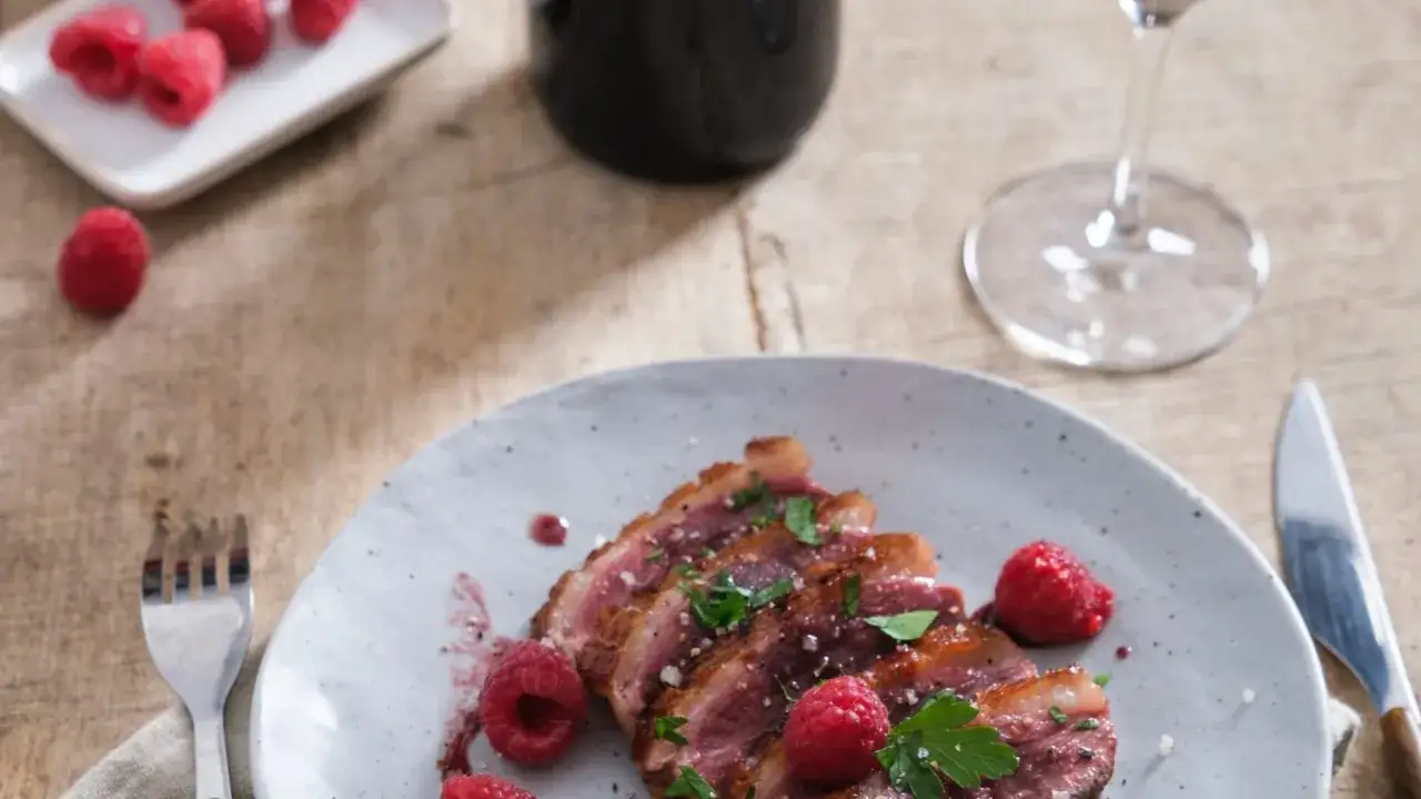 Tranches de magret de canard, garnies de framboises et persil, accompagnées d'un verre de vin rouge. Un délice pour les papilles.