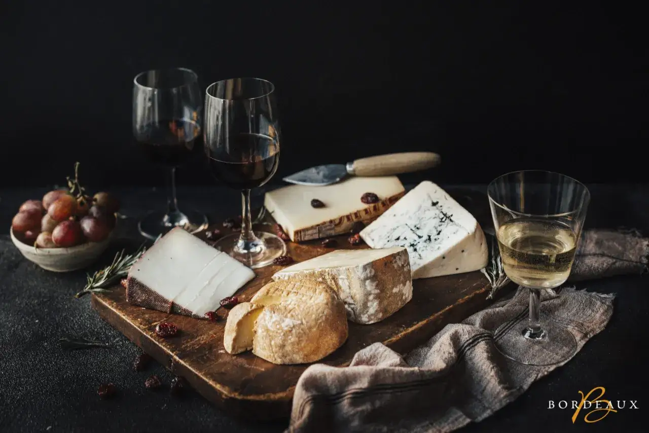 Un plateau de fromages variés avec des raisins, des airelles et des verres de vin rouge et blanc. Quel vin de Bordeaux choisir pour accompagner ce délice ?