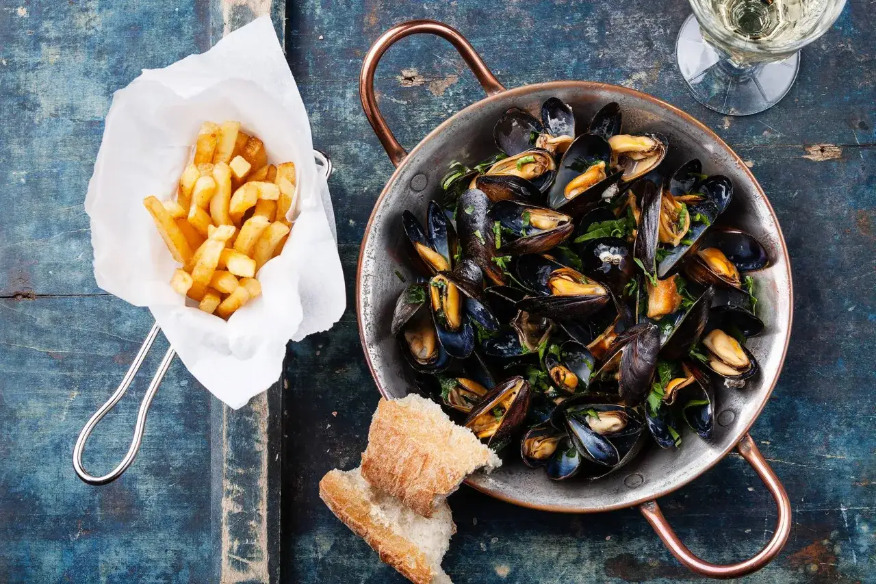 Un plat de moules frites, accompagné de pain et d'un verre de vin blanc, sur une table en bois bleu.