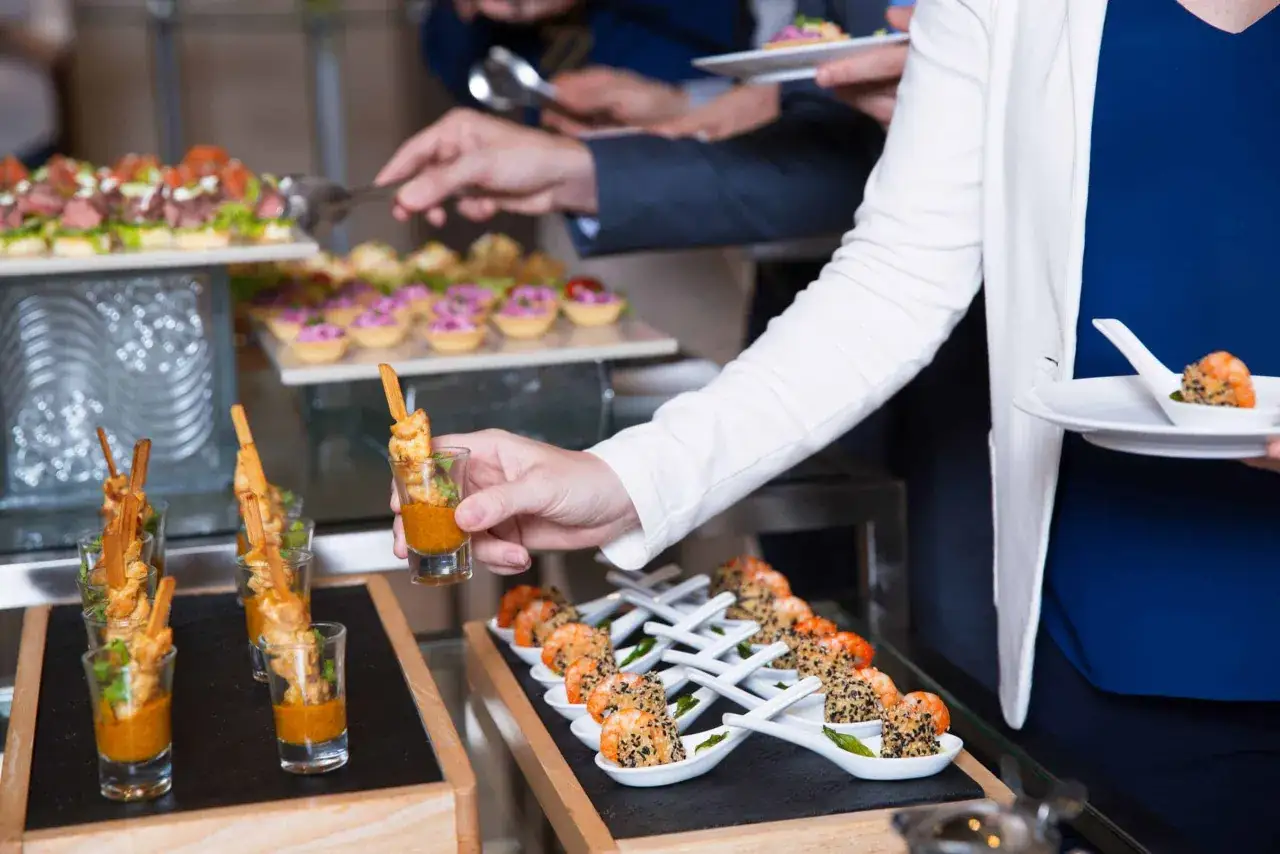 Un buffet restauration propose des verrines de crevettes et des brochettes. Une main saisit une verrine pour la déguster.