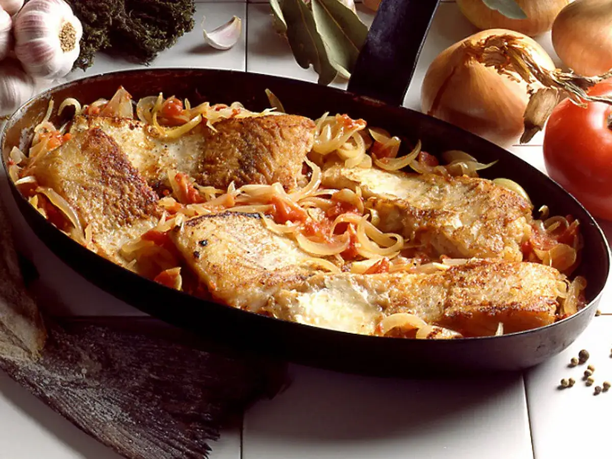 Morue &agrave; la lyonnaise, des filets de poisson dor&eacute;s mijotant avec des oignons et des tomates dans une po&ecirc;le.