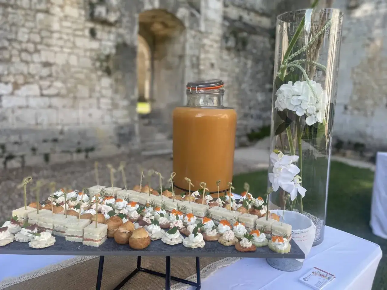 Un buffet de petits fours et sandwichs, idéal pour une boisson soirée, avec une grande carafe de jus orangé et des fleurs blanches dans un vase.
