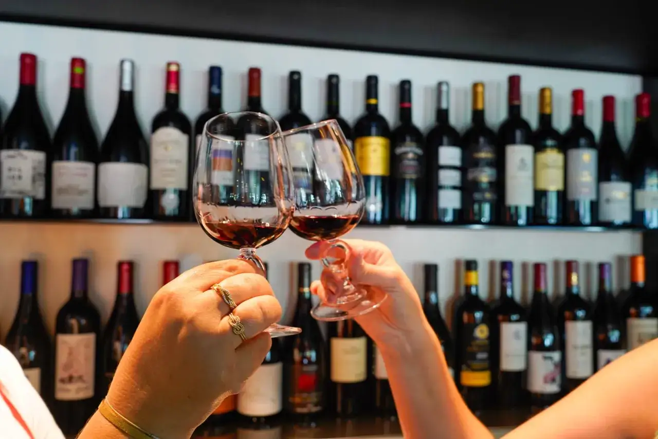 Deux mains portent des verres de vin rouge pour un toast, avec une étagère remplie de bouteilles, dont un millésime Cahors.