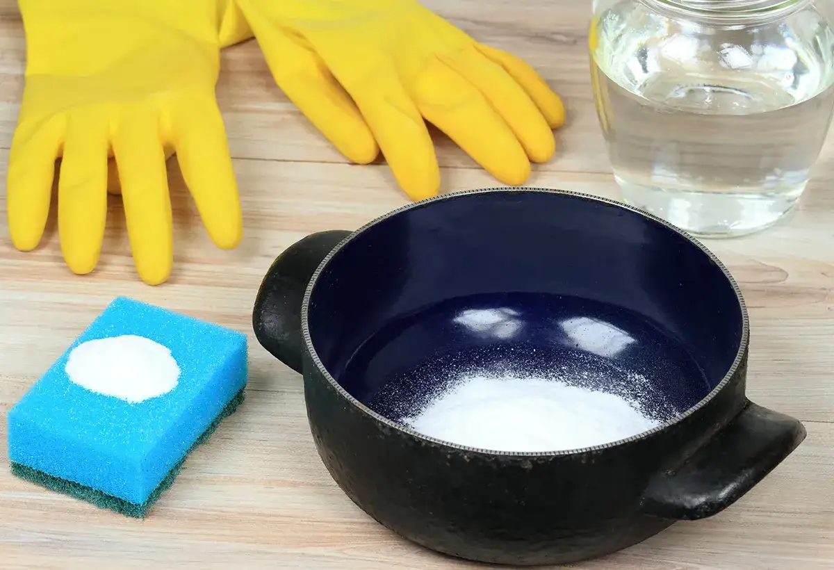 Gants jaunes, éponge bleue avec poudre blanche, casserole noire et bleue, et un bocal rempli de liquide clair. Idéal pour savoir comment enlever le cramer dans une casserole sans bicarbonate.