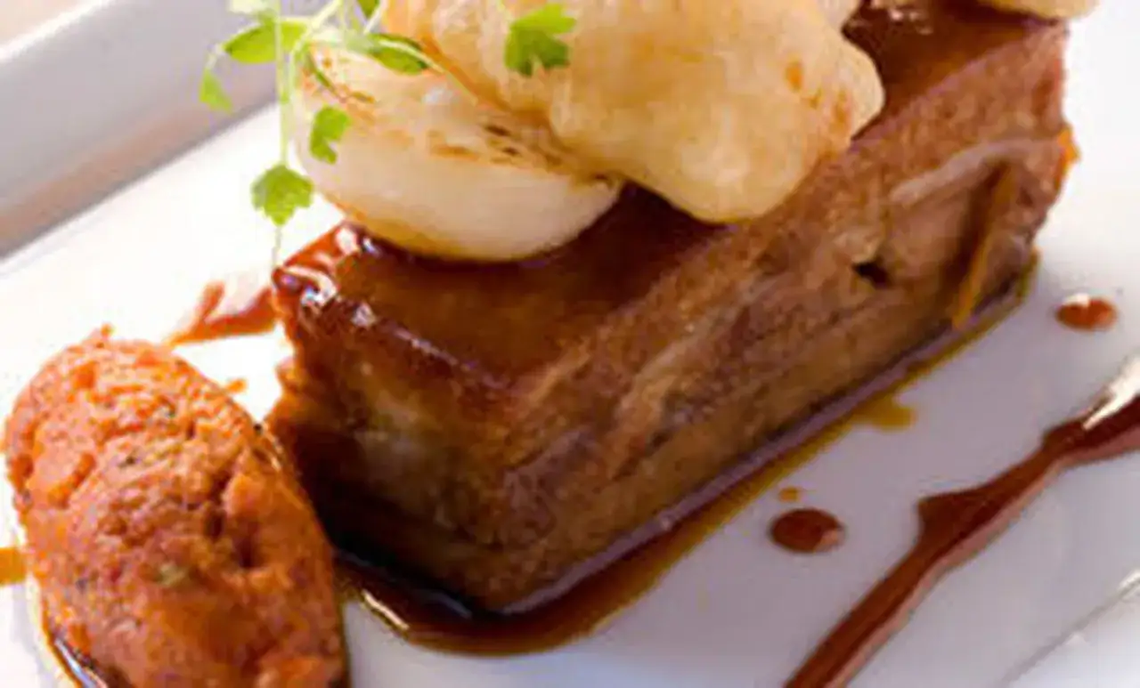 Un morceau de porc au caramel, nappé d'une sauce brillante, servi avec une purée orangée et garni d'oignons caramélisés et de persil.