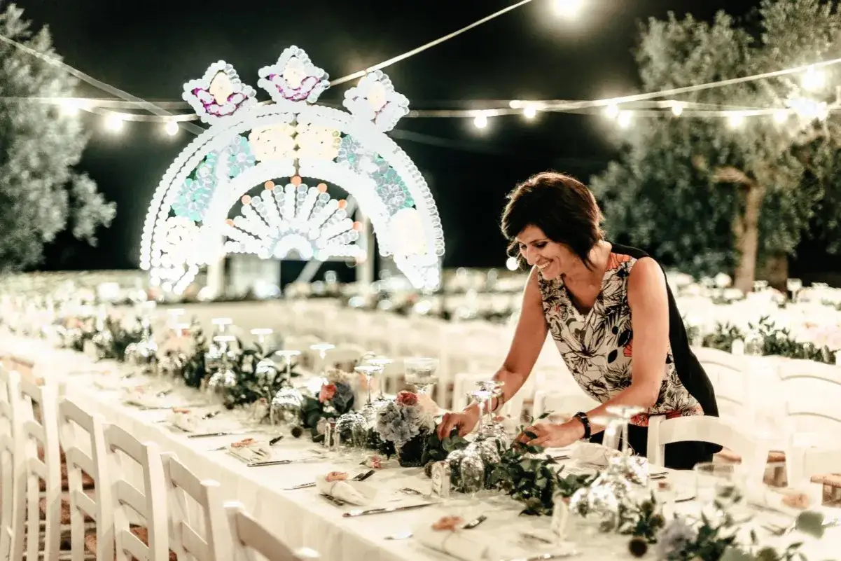 Une femme arrange des fleurs sur une table dress&eacute;e pour un mariage dolce vita, sous une arche lumineuse.