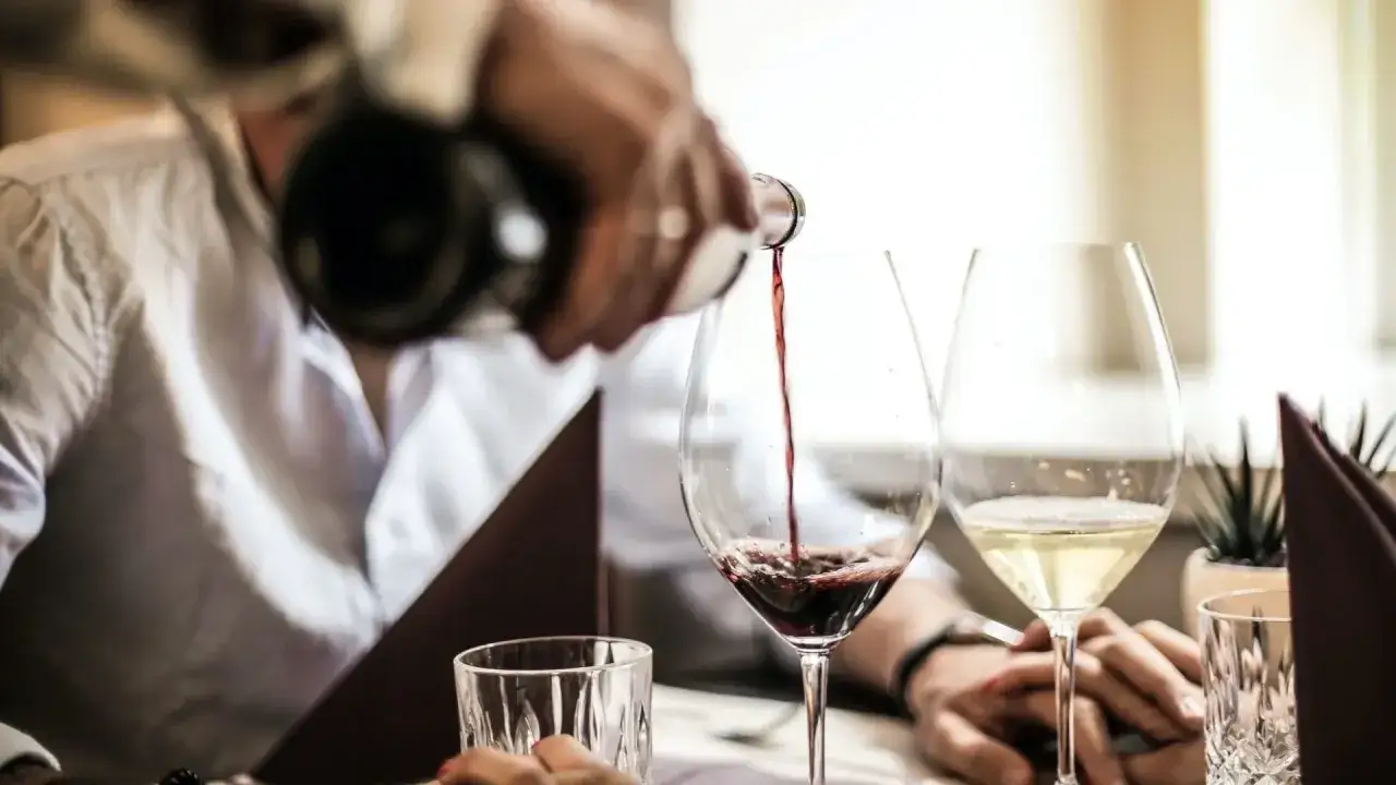 Un homme verse du vin rouge dans un verre, à côté d'un verre de vin blanc. Ambiance de bar feutrée.
