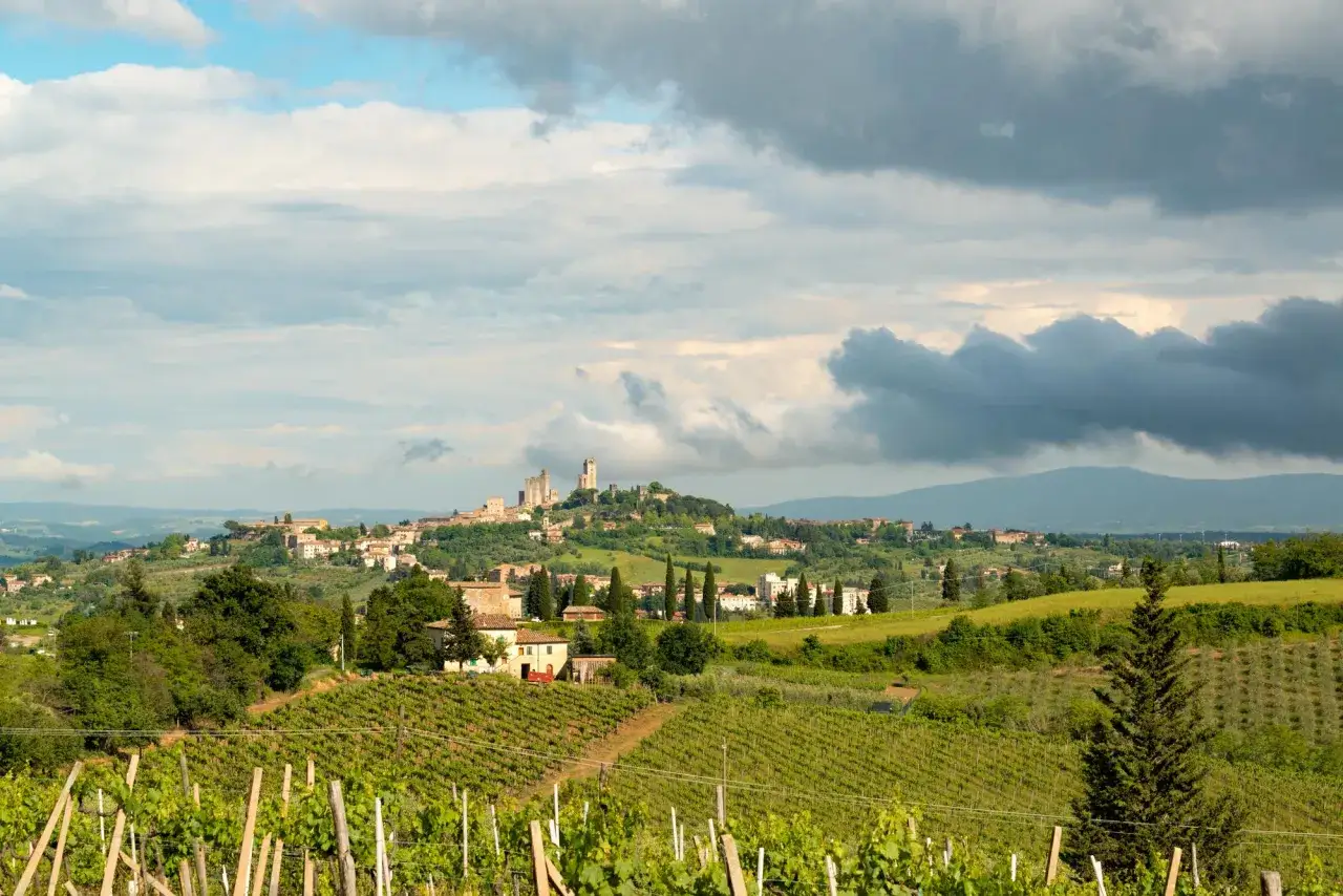 Paysage toscan avec vignobles verdoyants au premier plan, un village médiéval sur une colline et des nuages dramatiques. Un verre de chianti blanc serait parfait ici.