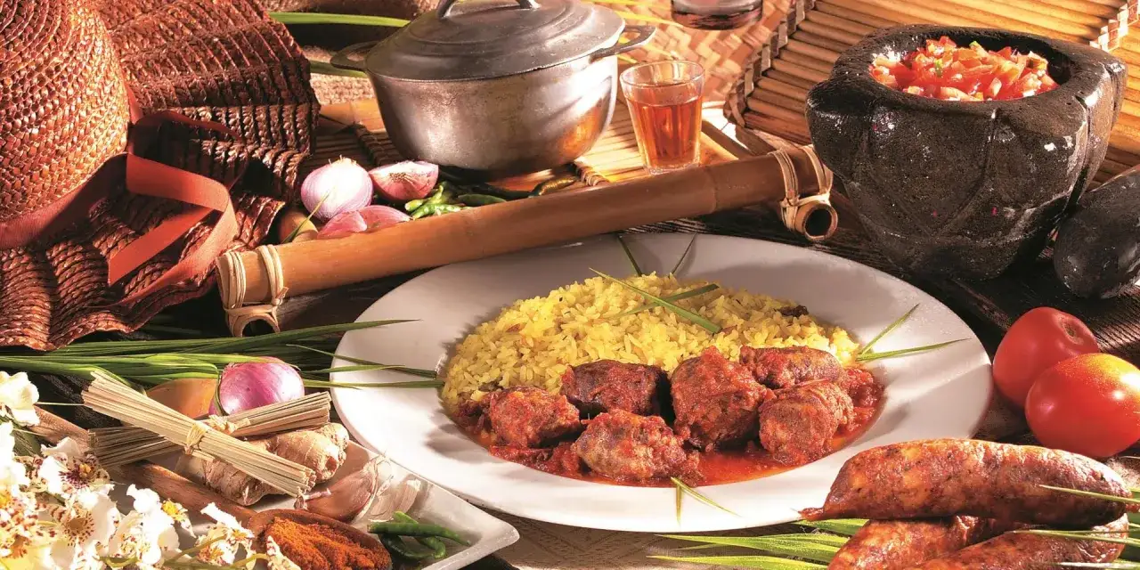 Un plat de riz jaune avec du rougail saucisse, accompagné de tomates, oignons, gingembre et épices. Un verre de rhum à côté.