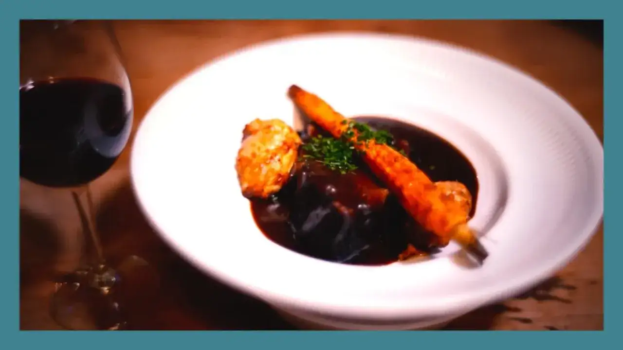 Un plat de paleron au vin rouge, tendre et savoureux, accompagné de carottes glacées et d'une touche de persil frais. Un verre de vin rouge complète ce tableau gourmand.
