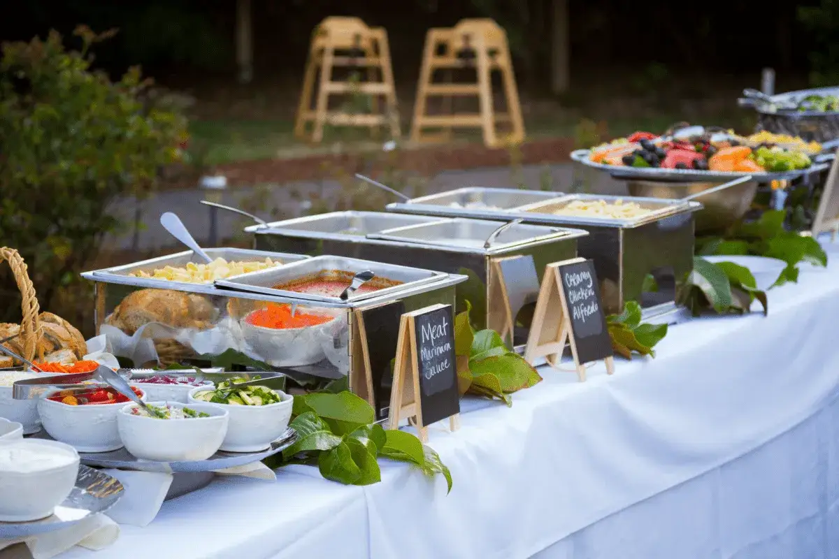 Buffet de mariage sans traiteur : plats chauds, salades et pains dispos&eacute;s sur une longue table blanche, d&eacute;cor&eacute;e de feuilles vertes.