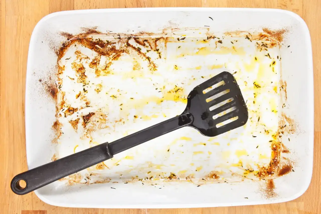 Une spatule noire repose dans un plat &agrave; four blanc sale, couvert de r&eacute;sidus br&ucirc;l&eacute;s et de graisse. Il est temps de nettoyer ce plat de four br&ucirc;l&eacute;.